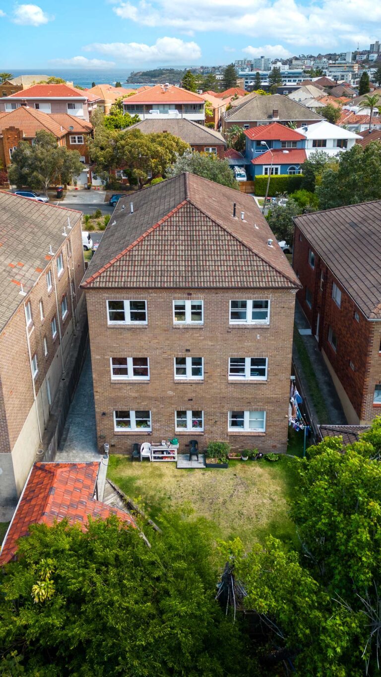 Aerial view of a three-story brick apartment building with a red-tiled roof, surrounded by similar buildings, green trees, and a grassy backyard on a clear day.