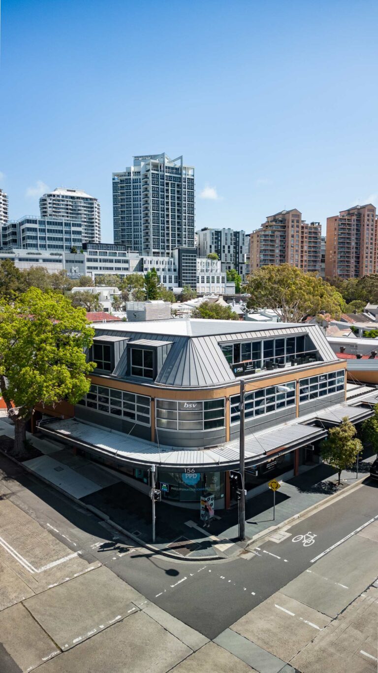 A two-story modern building on a street corner, with large windows and a metal roof, surrounded by trees and high-rise apartment buildings in the background under a clear blue sky.