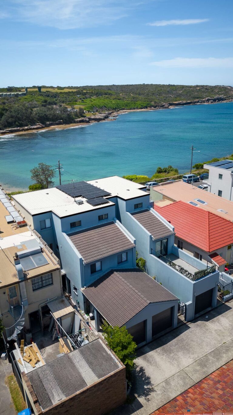 Aerial view of modern houses with gray and red roofs near a calm turquoise bay, with a green, tree-covered coastline in the background under a clear blue sky.