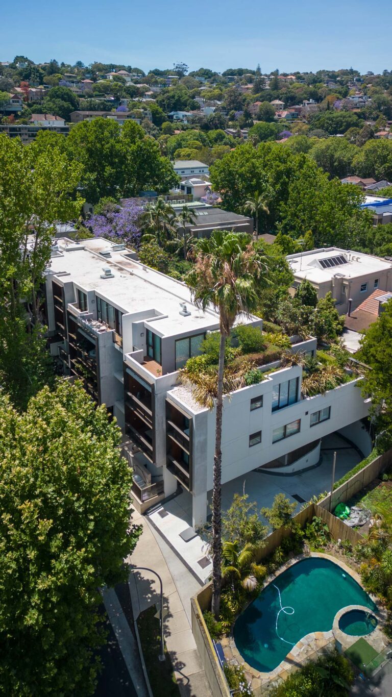 Aerial view of a modern apartment building with a green rooftop garden, surrounded by trees and residential homes. A kidney-shaped swimming pool is visible in the lower right corner.