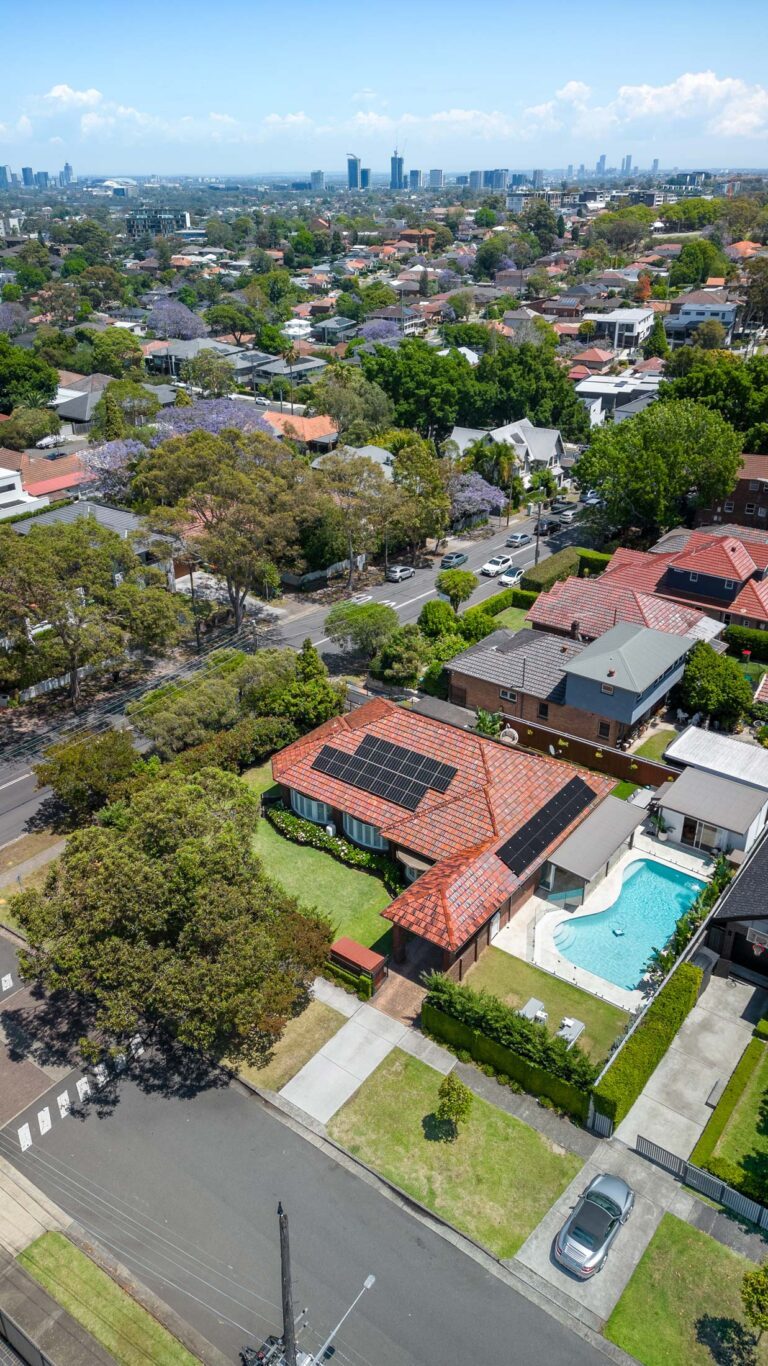 Aerial view of a suburban neighborhood with houses, trees, and a swimming pool. One house has solar panels on its red-tiled roof. City skyline is visible in the distant background under a partly cloudy sky.