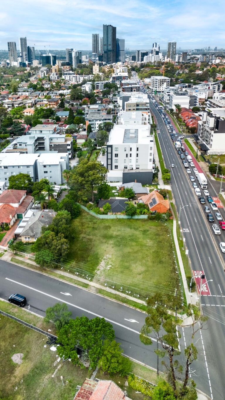 Aerial view of a cityscape with tall buildings in the background, residential houses, apartment buildings, green vacant lots, and busy streets with cars and a visible bus lane.