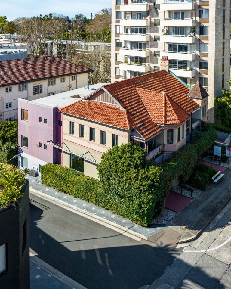 Aerial view of a corner house with a red tiled roof, surrounded by green hedges, next to a pink building and multi-story apartments on a sunny day.