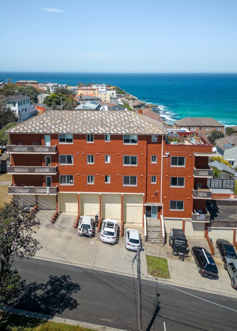 Aerial view of a red brick apartment building with balconies, several parked cars in front, and a coastal neighborhood leading to the turquoise ocean under a clear blue sky.