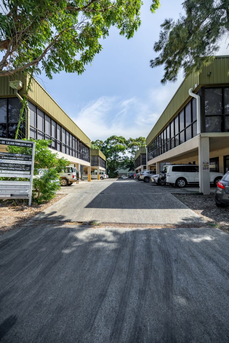 A paved driveway runs between two modern office buildings with large glass windows and parked cars. Trees and greenery surround the area under a partly cloudy sky.