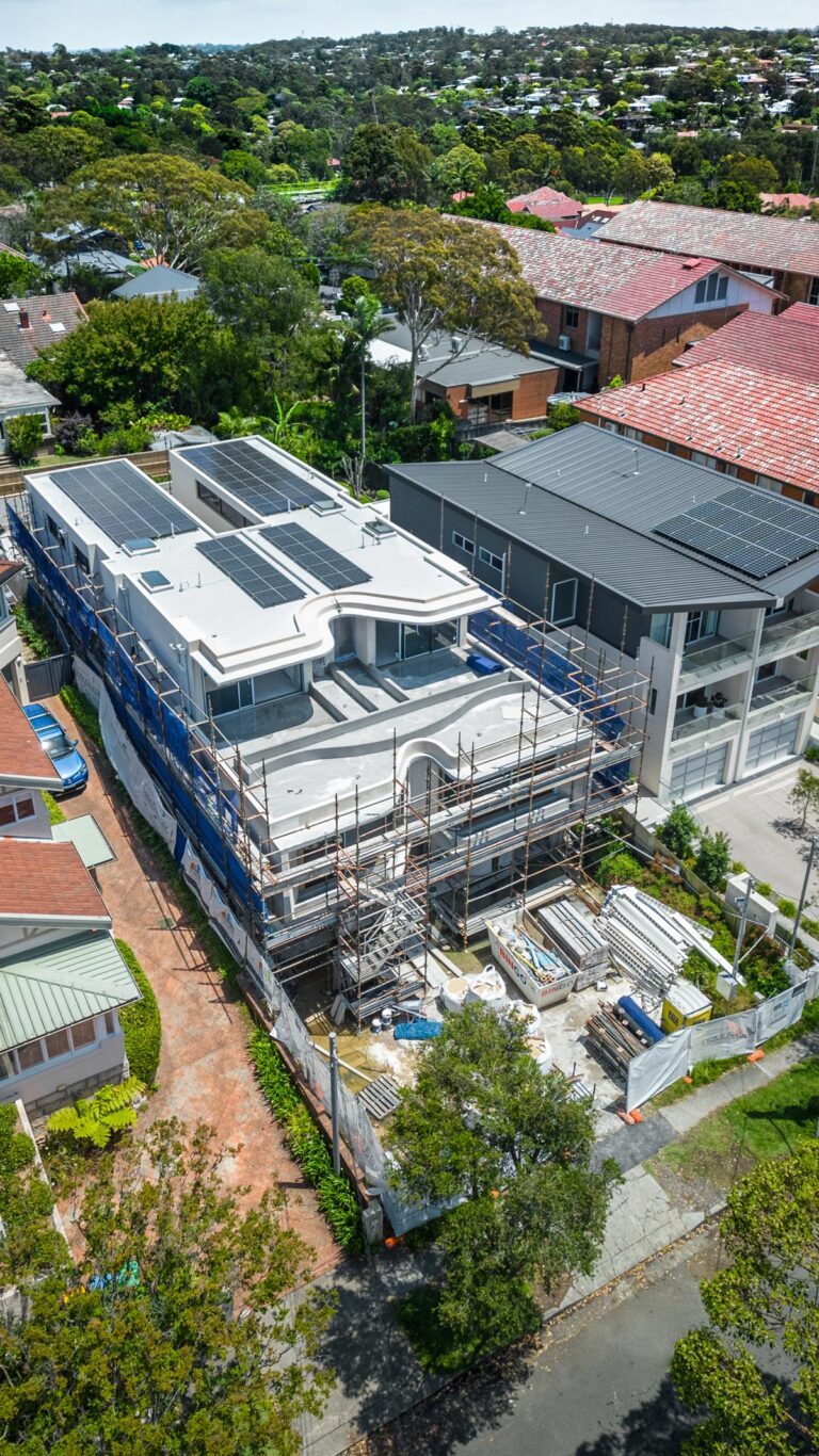 Aerial view of a large, modern house under construction with scaffolding, solar panels on the roof, and building materials stacked in the front yard, surrounded by other homes and trees.