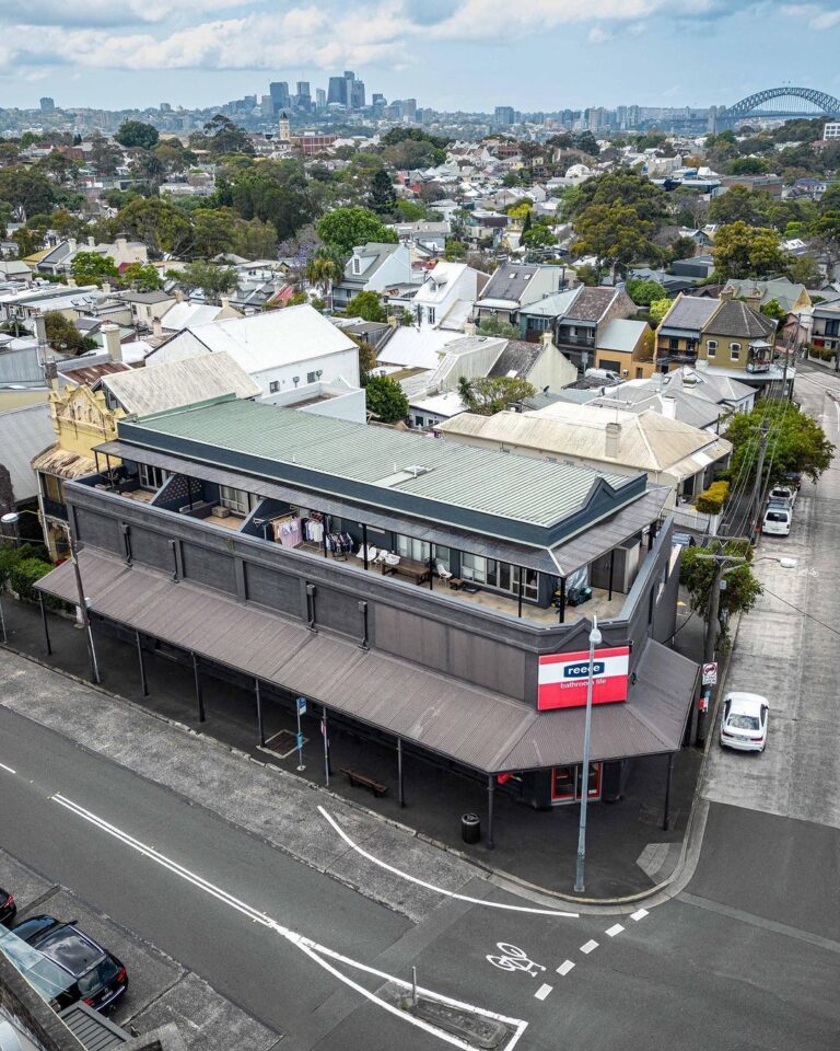 Aerial view of a large corner building with a wraparound awning, rooftop balcony, and red-and-white signage reading. Residential houses and Sydney city skyline, including the Harbour Bridge, are visible in the background.