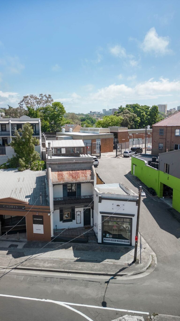 A street view shows two small, older buildings with weathered roofs, one white and one brown, next to a bright green wall. Behind them are more buildings, trees, and a blue sky with scattered clouds.