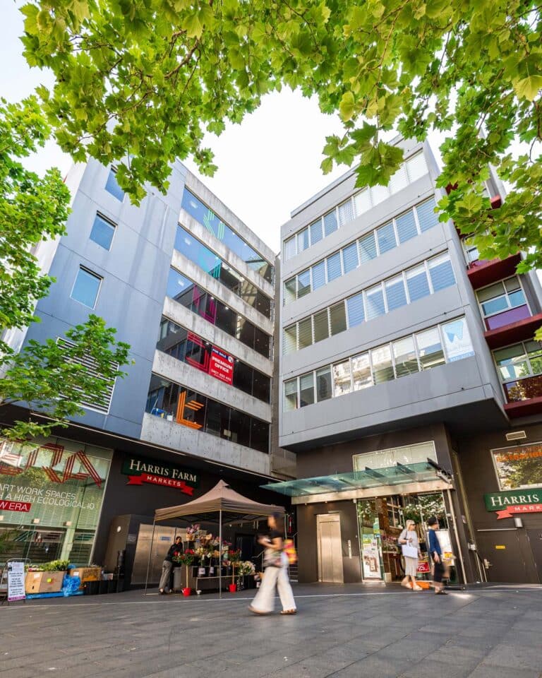 A street view shows modern apartment buildings with large windows, leafy green trees overhead, and people walking past a Harris Farm Markets store on the ground floor.