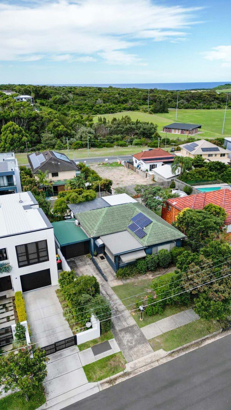 Aerial view of suburban houses with green roofs, solar panels, driveways, and gardens, bordered by a road in front and sports fields with trees and open sky in the background.