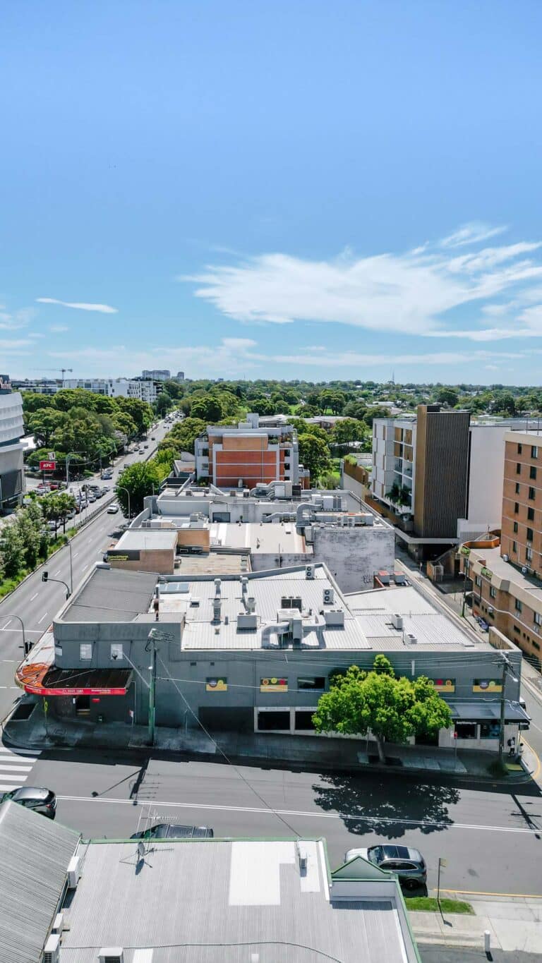 Aerial view of a city street lined with low-rise buildings, trees, and parked cars on a clear, sunny day with a blue sky and scattered clouds.