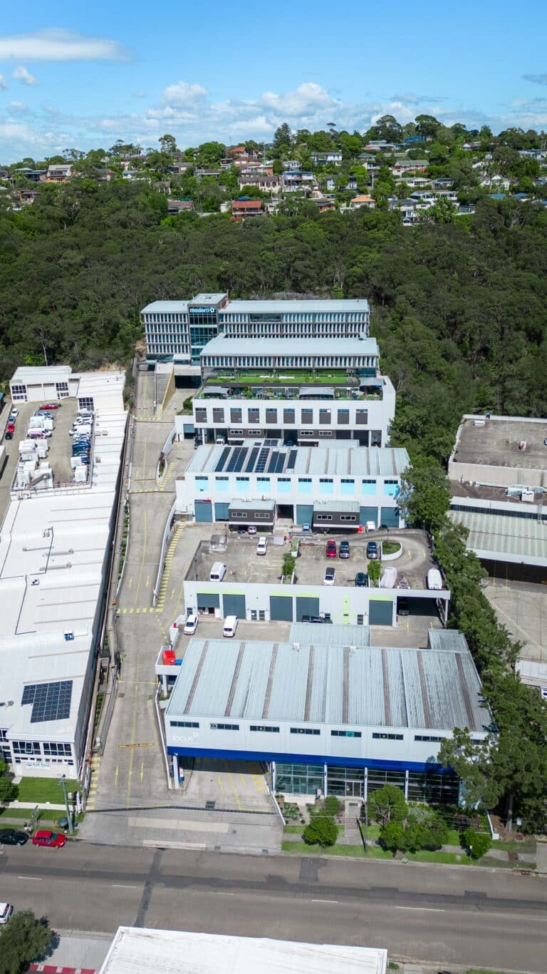 Aerial view of an industrial area with several blue and white buildings, parking lots with cars, and trees in the background. Residential houses are visible on a hill beyond the wooded area.