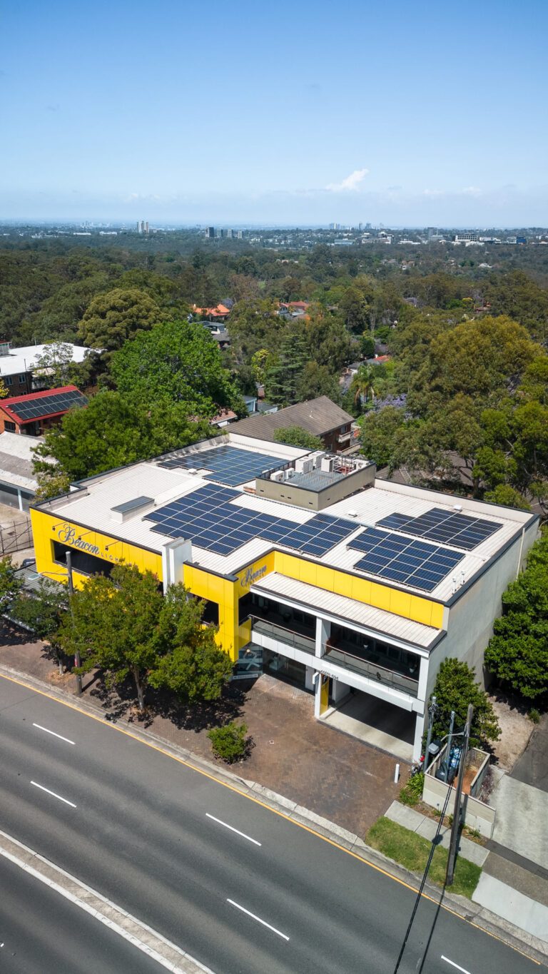 Aerial view of a modern, yellow and white commercial building with large solar panels on its roof, surrounded by trees and residential houses, adjacent to a main road under a clear blue sky.