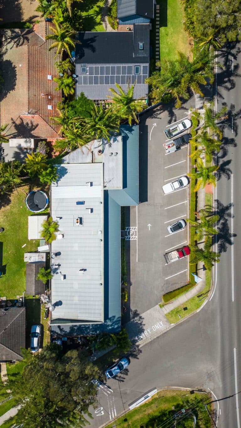 Aerial view of a building with a metal roof, surrounded by trees and parking lots with several parked cars; intersecting roads and crosswalks are visible on the right side of the image.