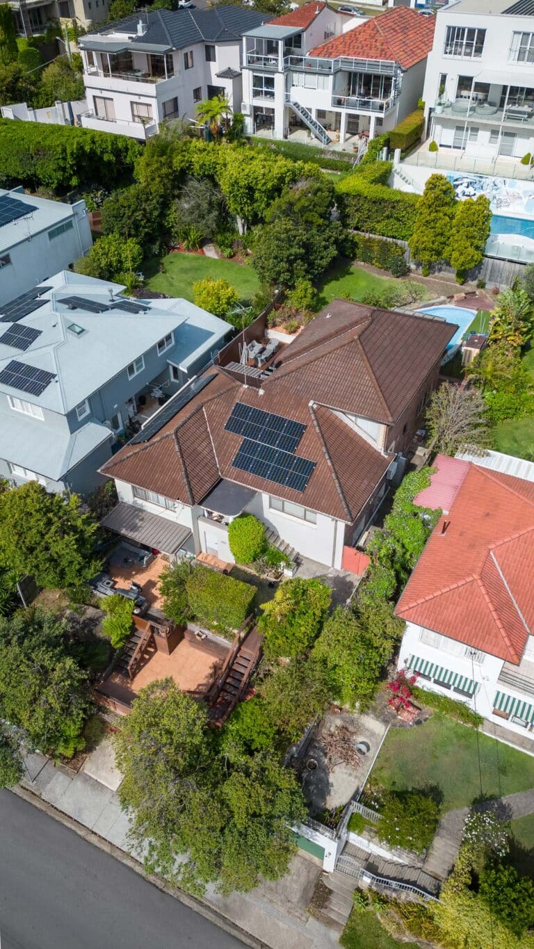 Aerial view of a suburban neighborhood featuring a house with solar panels on its brown, tiled roof, surrounded by greenery, other houses, and a neatly landscaped front yard.