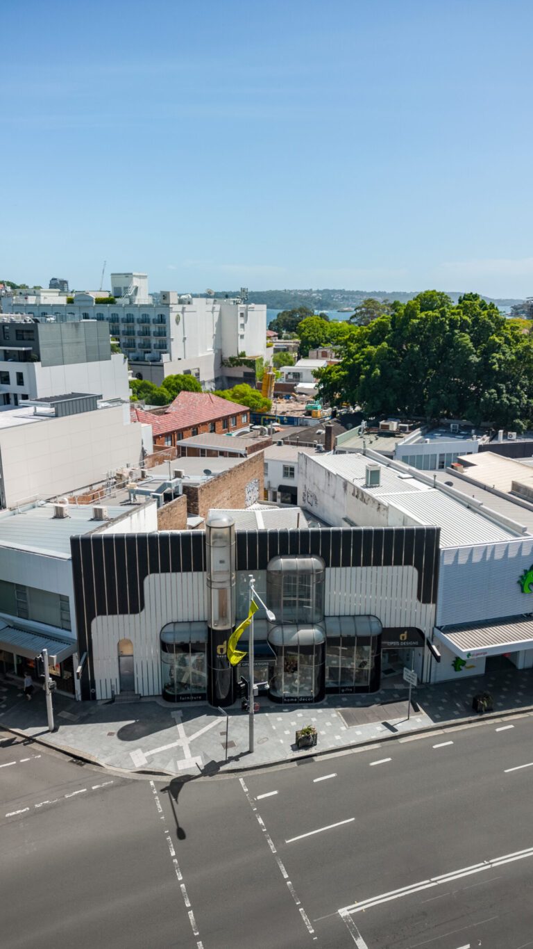 Aerial view of a city street corner with modern low-rise buildings, large windows, some greenery, and clear blue sky in the background. Crossing lines and street signs are visible.