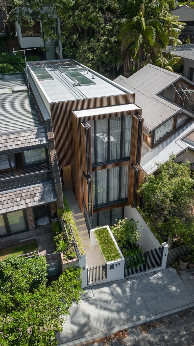 A modern, narrow three-story house with large glass windows and wooden slat accents, surrounded by greenery and neighboring homes, viewed from above. A small garden and a black metal gate are at the entrance.