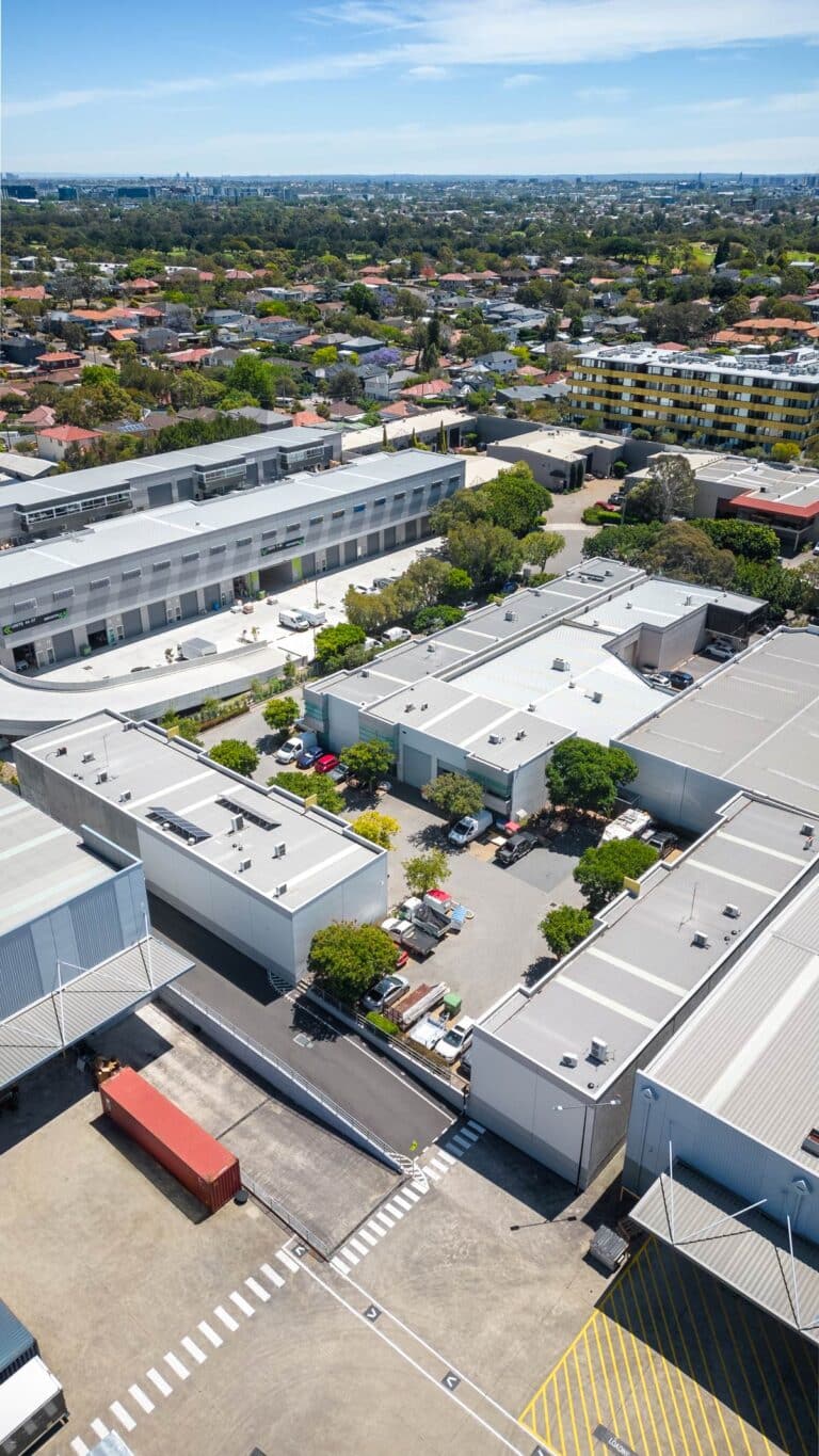 Aerial view of an industrial complex with white-roofed warehouses, surrounded by trees and parked vehicles, in a suburban area with houses and buildings in the background under a clear blue sky.