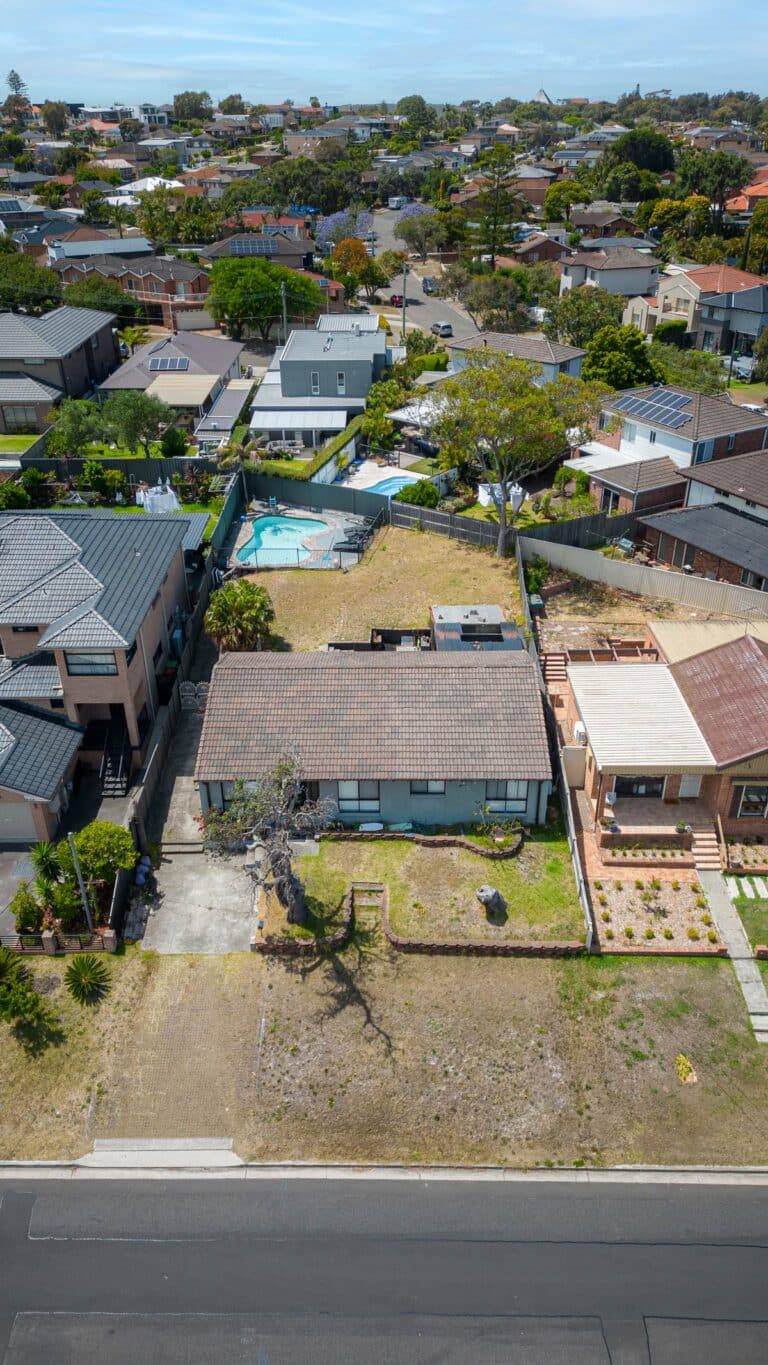 Aerial view of a suburban neighborhood showing houses with backyards, one with a pool, and front lawns along a quiet street on a sunny day.