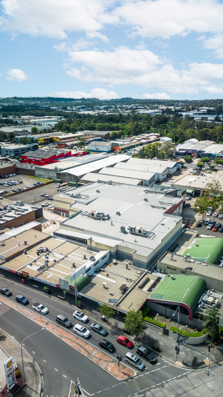 Aerial view of a commercial area with several large warehouses and retail stores, parking lots with cars, and a busy intersection in the foreground. Hills and partly cloudy sky can be seen in the background.
