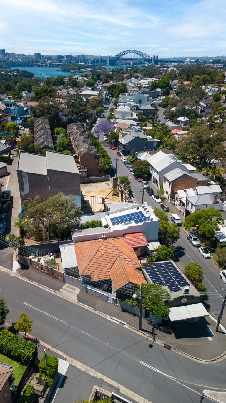 Aerial view of a suburban neighborhood with residential houses, some with solar panels on rooftops, tree-lined streets, and the Sydney Harbour Bridge visible in the distance under a partly cloudy sky.