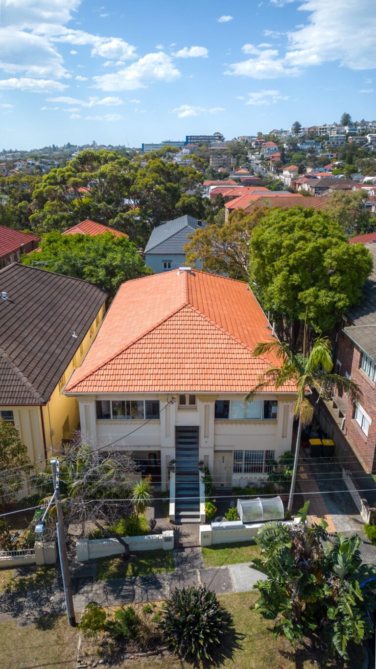 Aerial view of a two-story house with a red-tiled roof, white exterior, and a central front stairway, situated between neighboring homes with trees and greenery in a suburban neighborhood.