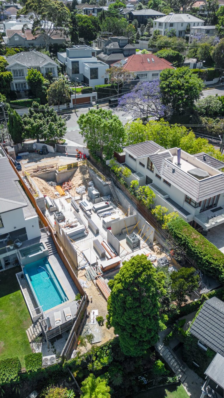 Aerial view of a residential neighborhood with a large house under construction, surrounded by completed homes, trees, and a swimming pool in an adjacent yard, on a sunny day.