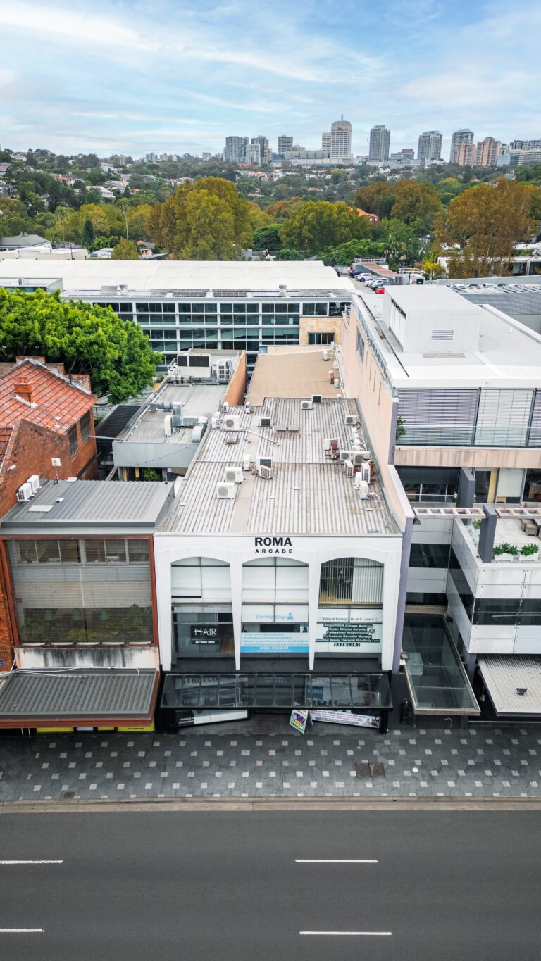 Aerial view of a white commercial building labelled “ROMA” on a city street, surrounded by other buildings, with trees and tall city buildings in the background under a blue sky.