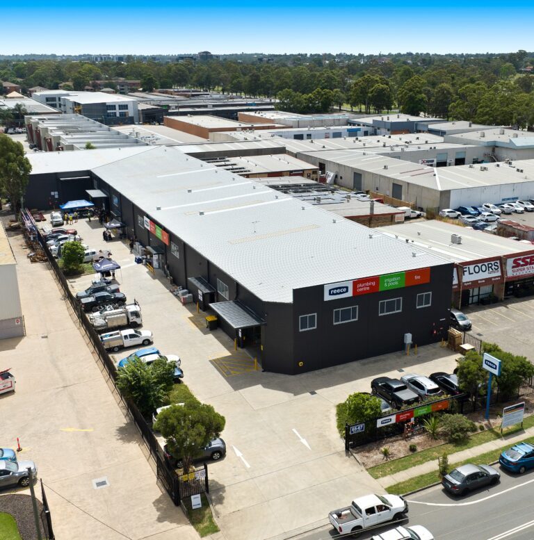 Aerial view of a modern industrial complex with multiple warehouses, parked vehicles, and a main entrance. The buildings have grey roofs and various business signs. Trees and other industrial buildings are in the background.