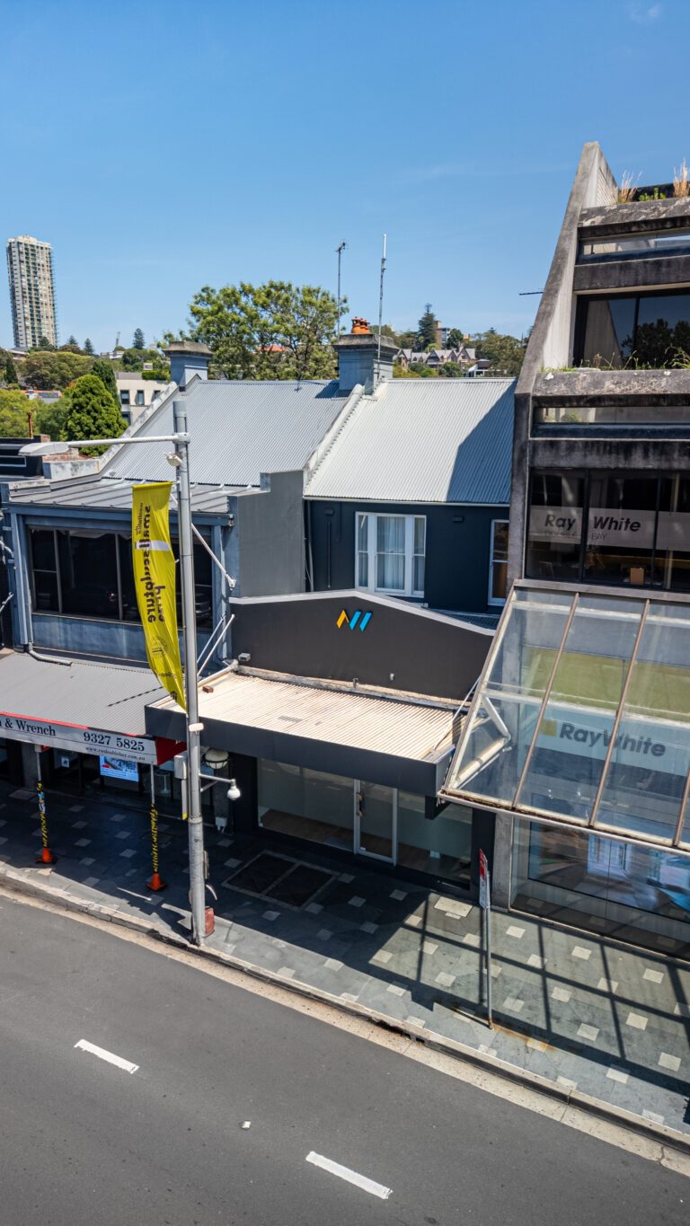 A city street view showing storefronts, including a gray building with a geometric logo, a yellow flag reading Auction, and a Ray White real estate office. Trees and a tall building appear in the background under a clear sky.