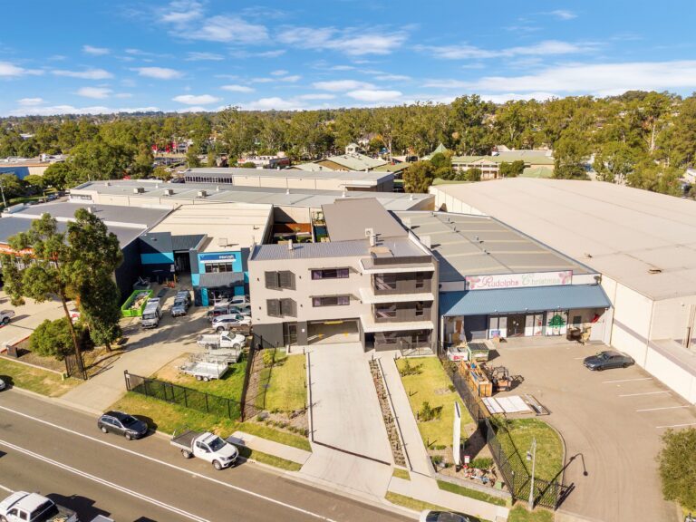 Aerial view of a modern industrial complex with multiple warehouse buildings, a driveway, parked cars, and trees in the background under a clear blue sky.