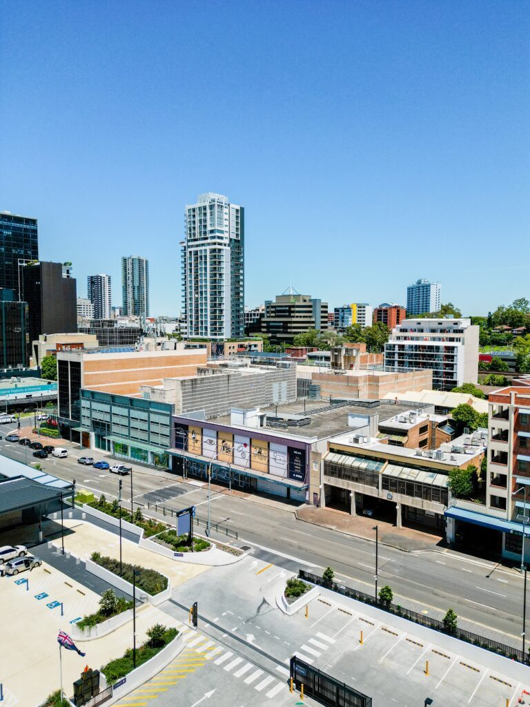 Aerial view of a modern cityscape with high-rise buildings, shops, and parking areas under a clear blue sky. Streets intersect in the foreground and few cars are visible.