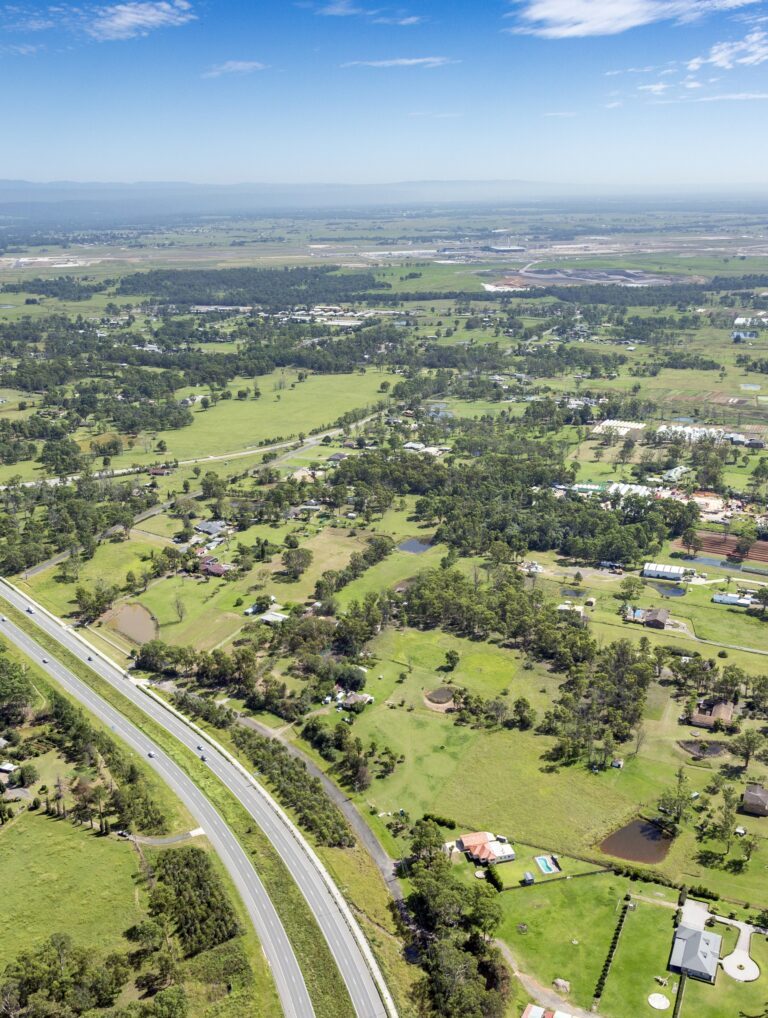 Aerial view of a rural area with green fields, scattered houses, trees, a main road curving through the landscape, and distant mountains under a partly cloudy blue sky.