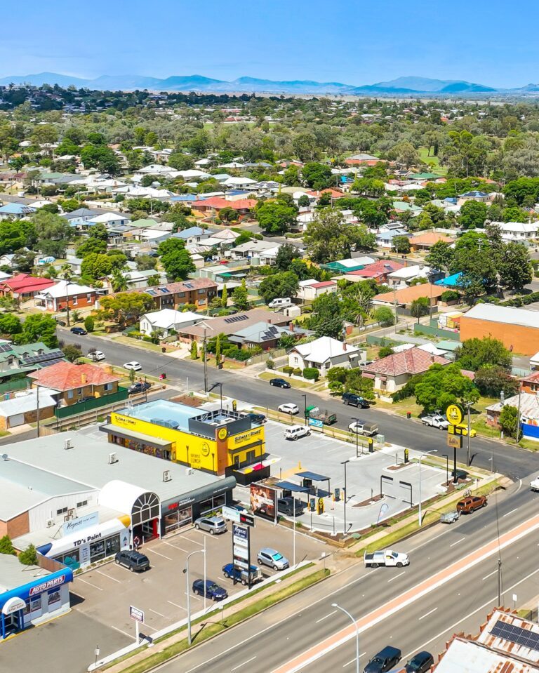 Aerial view of a suburban area with houses, trees, and streets; a yellow fast-food restaurant and several businesses are visible along a main road, with distant hills under a clear blue sky.