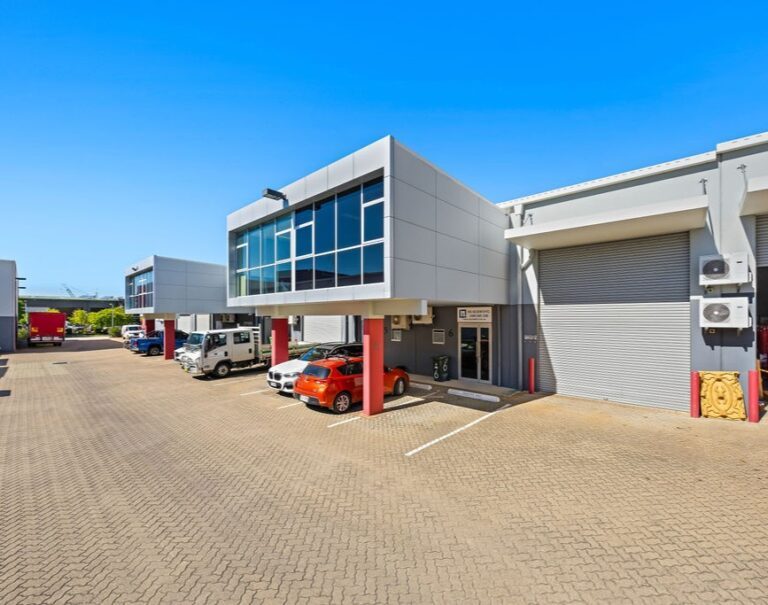 A modern commercial building with large glass windows, red pillars, and a roller door, surrounded by parked vehicles in a paved lot under a clear blue sky.