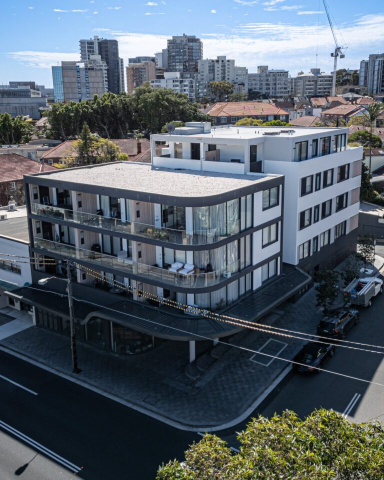 A modern, multi-story apartment building with glass balconies on a street corner, surrounded by cars, trees, and other residential buildings, with city high-rises and a crane visible in the background.
