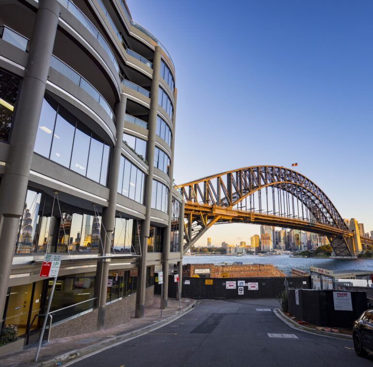A curved office building stands near the Sydney Harbour Bridge at sunset, with the city skyline and harbor visible in the background under a clear blue sky.