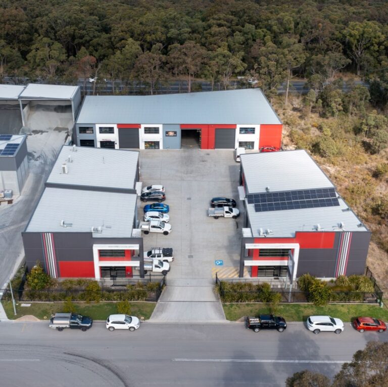 Aerial view of two modern commercial buildings with red and gray facades, solar panels on one roof, cars parked in front and in the central driveway, surrounded by trees and a forested area in the background.