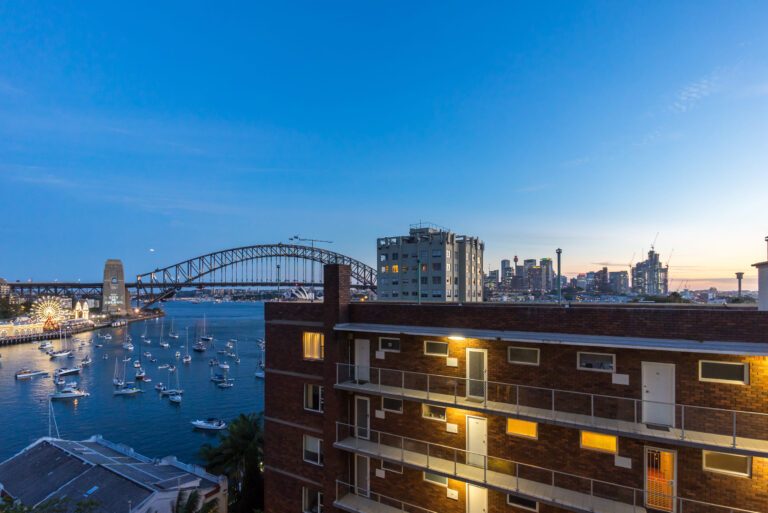 View of Sydney Harbour at dusk, featuring the Sydney Harbour Bridge, numerous boats on the water, city skyline in the background, and a lit brick apartment building in the foreground.