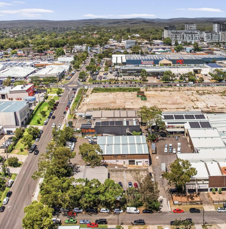 Aerial view of a suburban commercial area with warehouses, parking lots, parked cars, shopping centers, and surrounding trees under a partly cloudy sky. Residential and commercial buildings are visible in the distance.