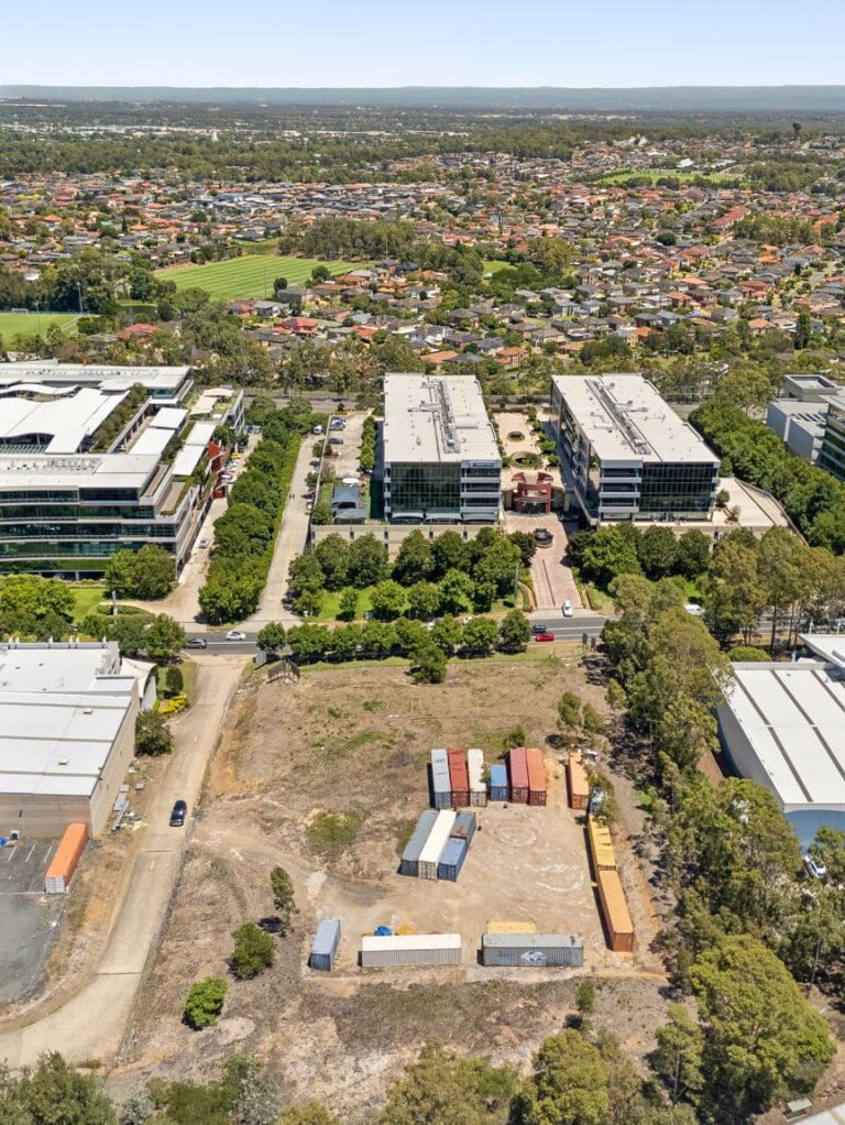 Aerial view of an urban area with office buildings, a cleared lot containing shipping containers, tree-lined streets, and residential neighborhoods in the background.