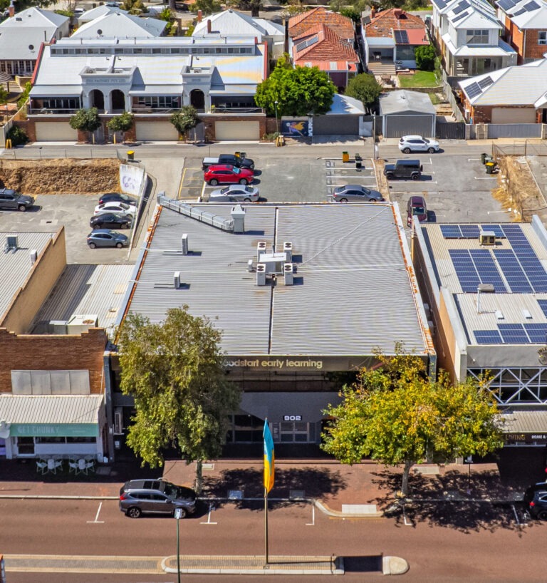 Aerial view of a childcare center, labeled Goodstart early learning, with parked cars in front and behind, surrounded by residential houses and other buildings. A tree and a flag are visible on the sidewalk.