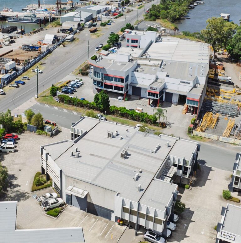 Aerial view of an industrial area with several large warehouse buildings, parked cars, and construction materials. A river and more industrial facilities are visible in the background under a clear sky.