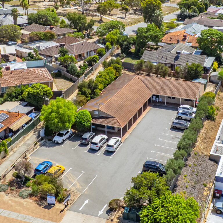 Aerial view of a single-story brick building with a tiled roof and a small parking lot, surrounded by trees and residential houses in a suburban neighborhood. Several cars are parked in the lot.