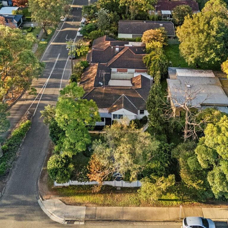 Aerial view of a suburban neighborhood showing houses with tiled roofs, surrounded by trees and greenery, bordered by two intersecting streets.