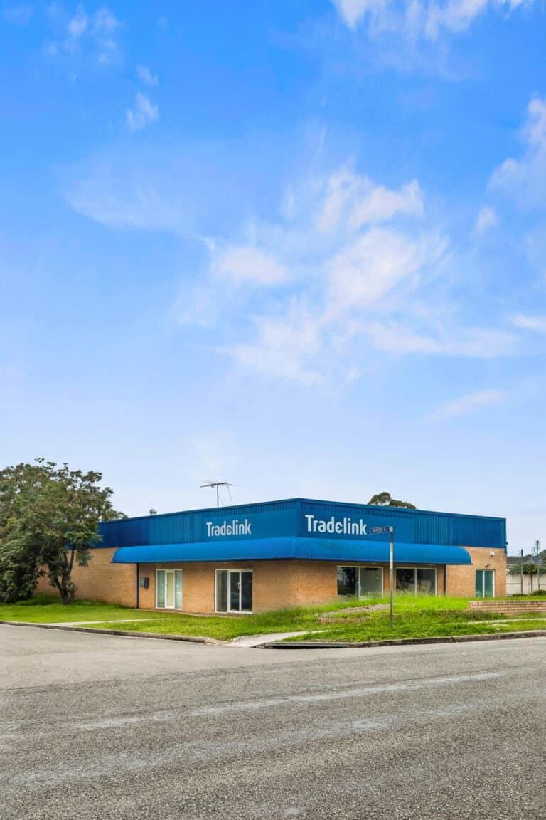 A single-story commercial building with a blue roof and Tradelink signage on the front, located on a corner lot with a street and greenery surrounding it under a clear blue sky.