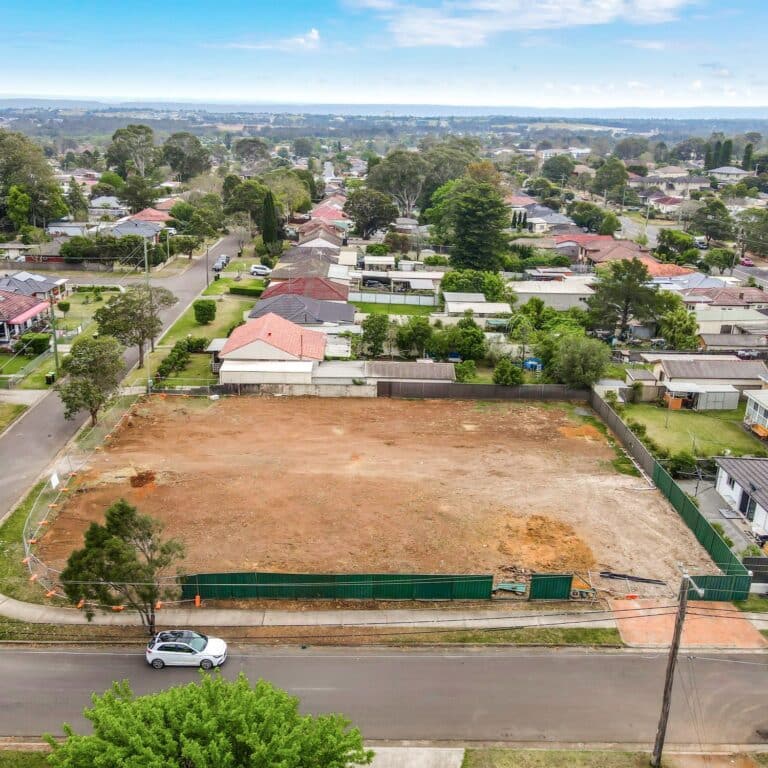 A vacant, leveled dirt lot is bordered by green fencing in a suburban neighborhood, with houses and trees lining the surrounding streets under a partly cloudy sky. A single white car is parked on the street in front.