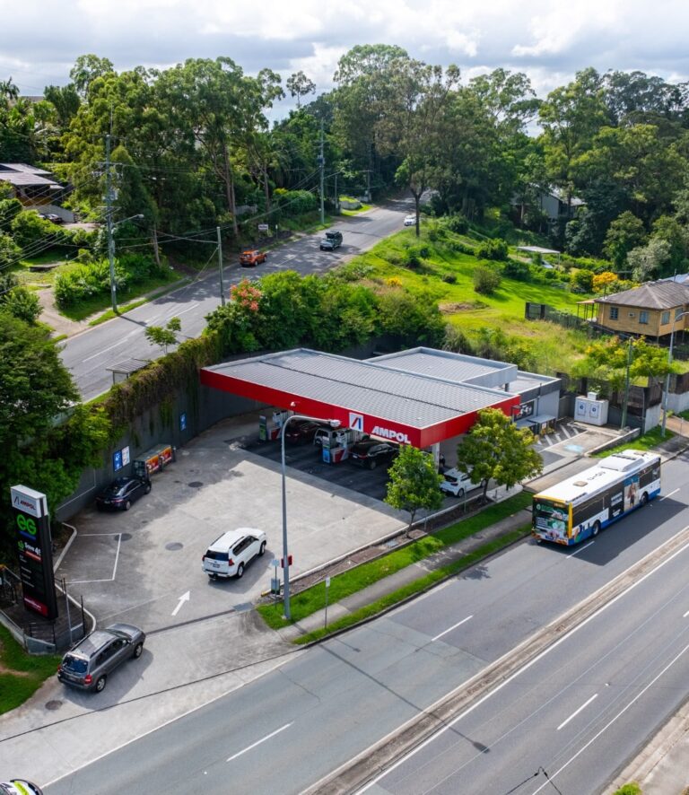 An aerial view of an Ampol gas station with several cars parked at pumps, a bus passing by on the main road, and green trees and houses in the background.