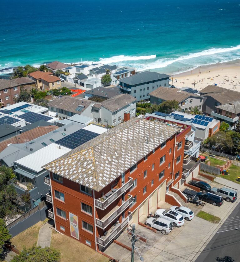 Aerial view of a coastal neighborhood with a red brick apartment building in the foreground, cars parked below, and a sandy beach with turquoise ocean waves in the background.