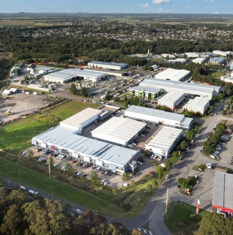 Aerial view of an industrial estate with multiple large white-roofed warehouse buildings, surrounded by roads, cars, green spaces, and distant residential areas under a partly cloudy sky.